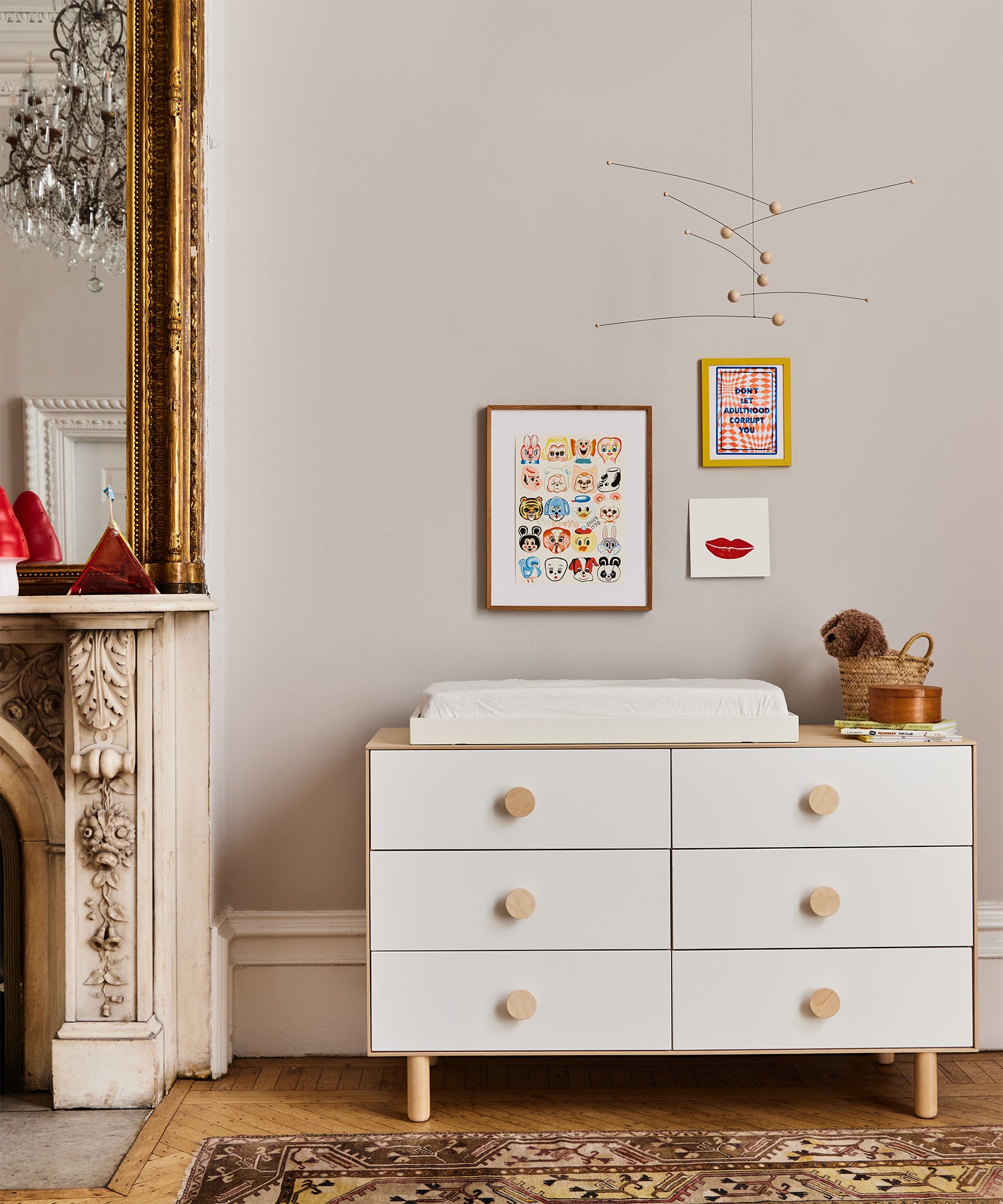 Modern nursery with white dresser, changing tray, wall art, mobile, and an ornate fireplace topped with a mirror.