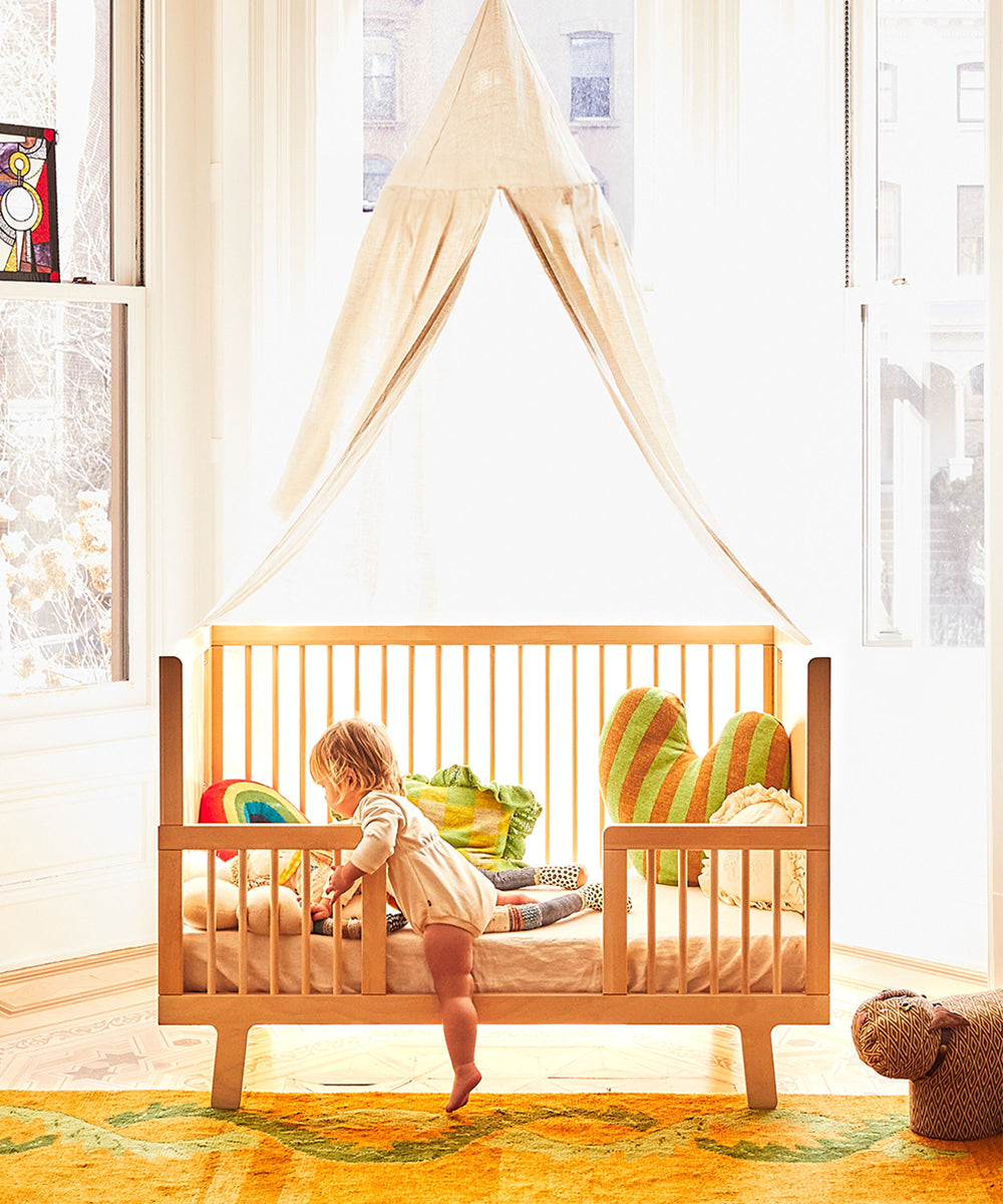 A toddler stands on a Dual Firm Crib Mattress in a wooden crib with colorful pillows, beneath a canopy in a sunlit room.