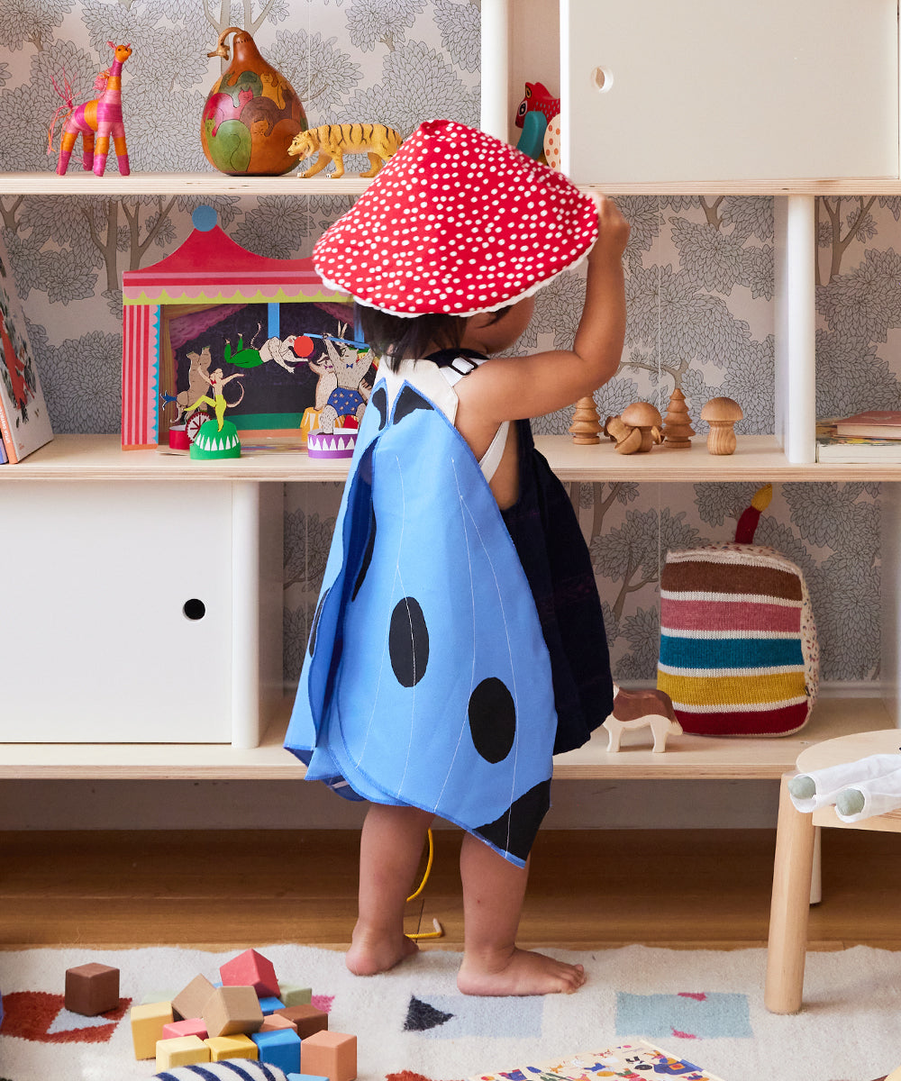 A toddler in a red mushroom hat and Le Blue Butterfly Wings Costume plays in a colorful, toy-filled room with shelves and blocks.