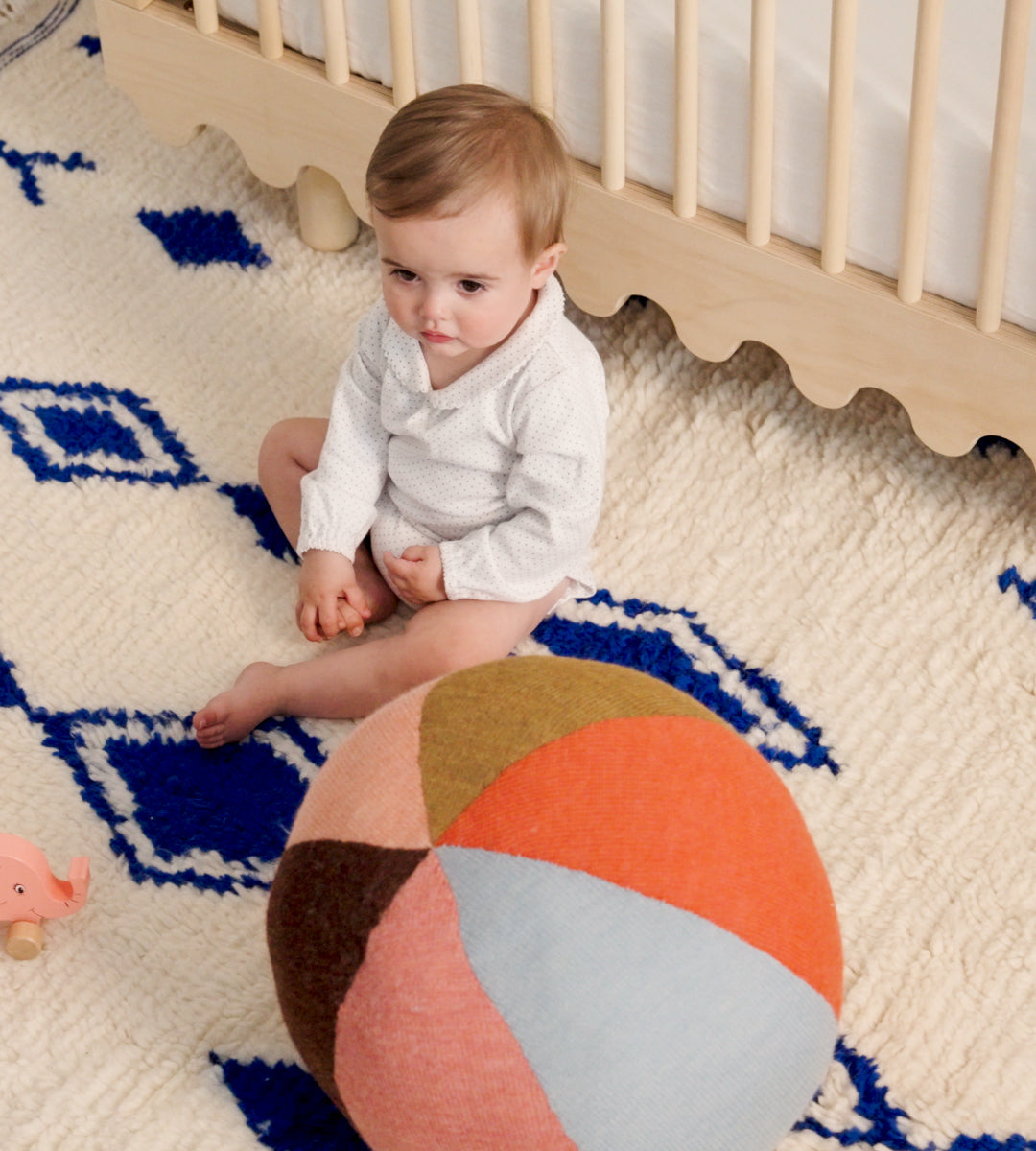 Baby sitting on a patterned rug near a crib, with the Circus Ball - Rose colorful round cushion and a small toy nearby.