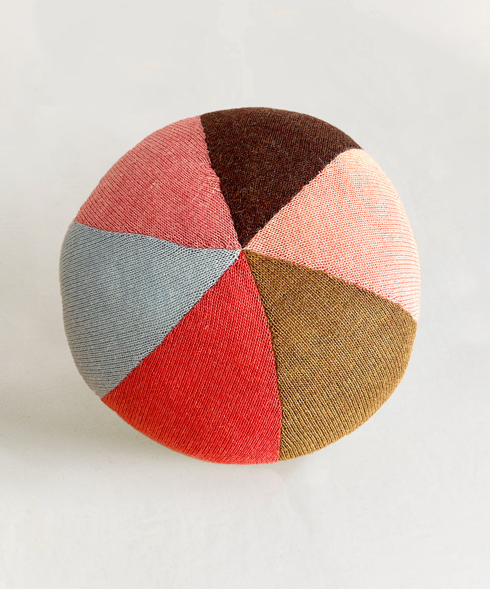 Circus Ball - Rose: A round pillow with triangular sections in red, brown, pink, yellow, and blue on a light background.