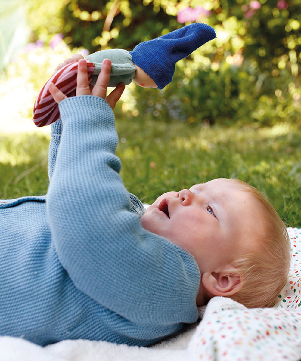 A baby lying on a blanket outdoors holds up the green Clown Rattle and examines it closely.