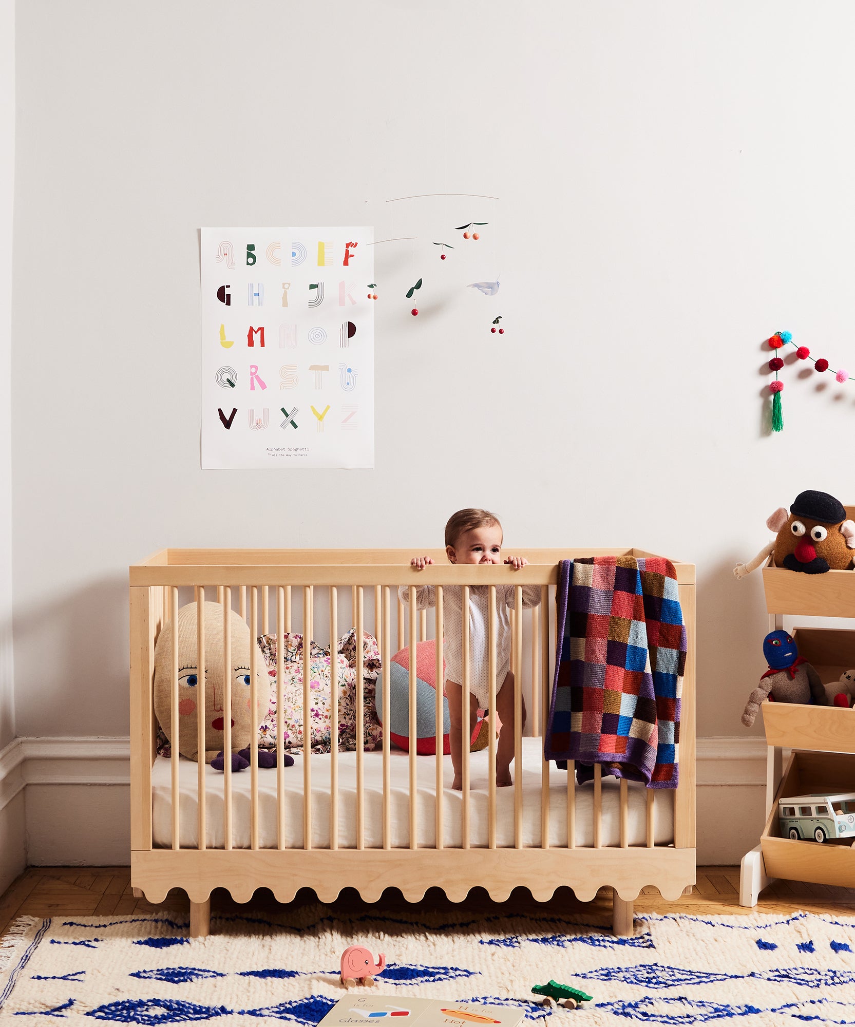 A baby stands in a wooden crib with a Patchwork Blanket, surrounded by colorful toys and decor in a cozy, modern nursery.
