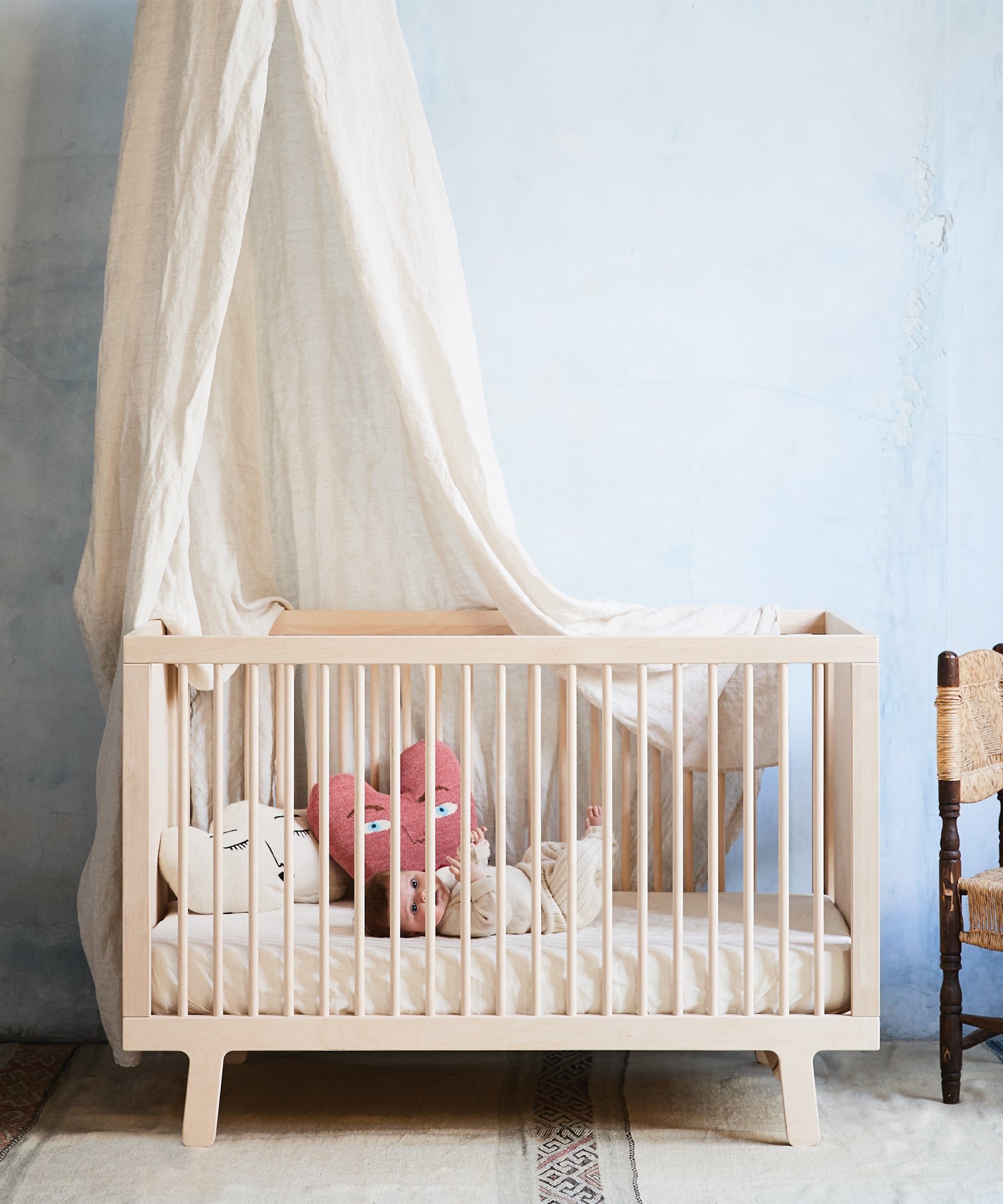 A baby rests in a crib dressed with a Linen Crib Sheet, under a white canopy, set against a light blue wall.