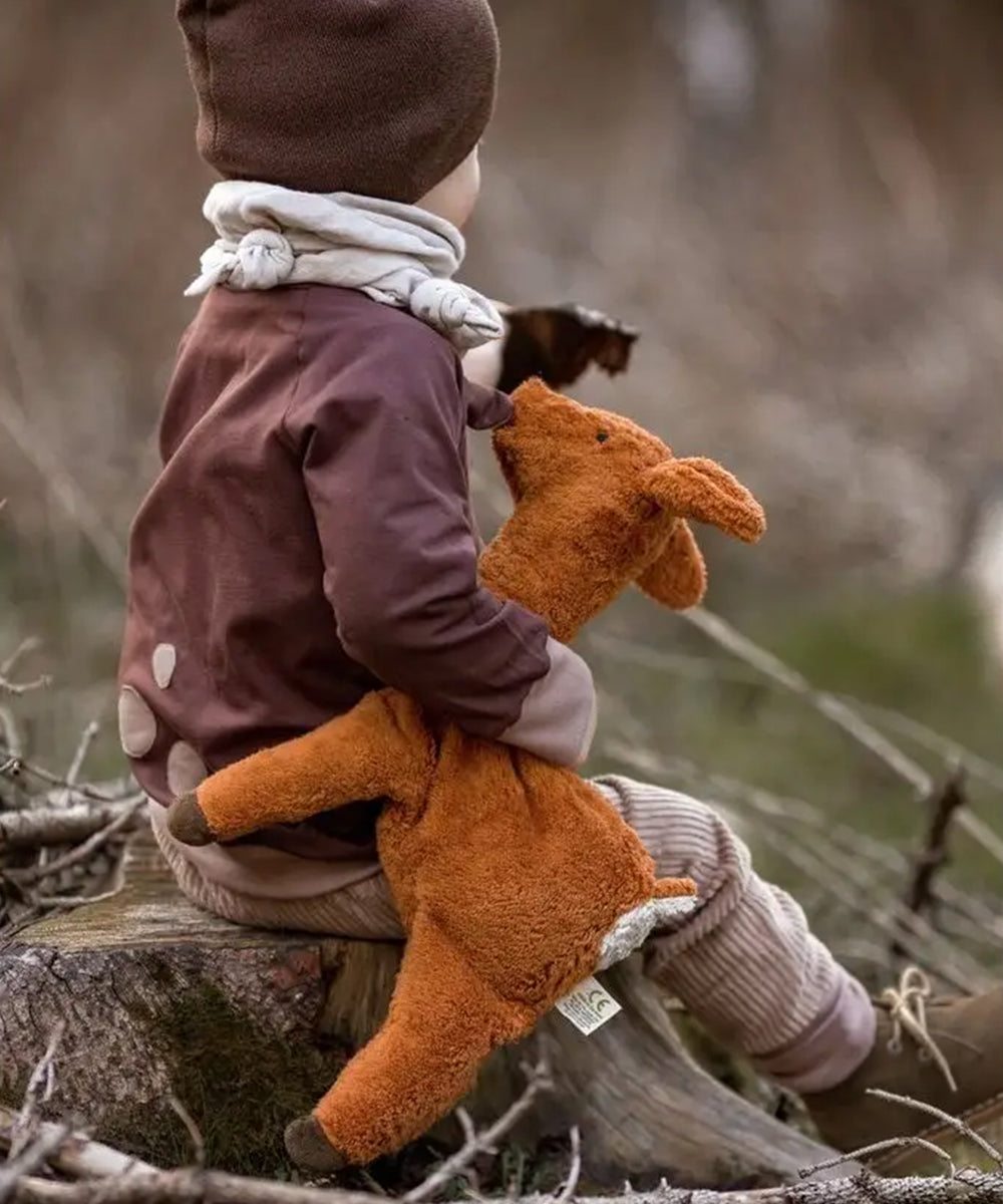 Child in brown clothes sits on a log outdoors, holding a plush orange deer toy and facing away.