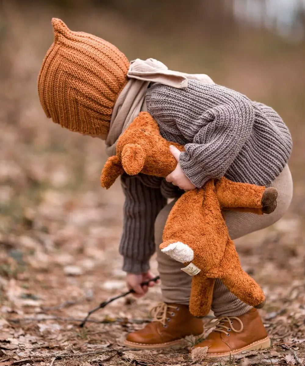 A child in cozy autumn clothes holding a plush deer bends down to pick up a stick in the woods.