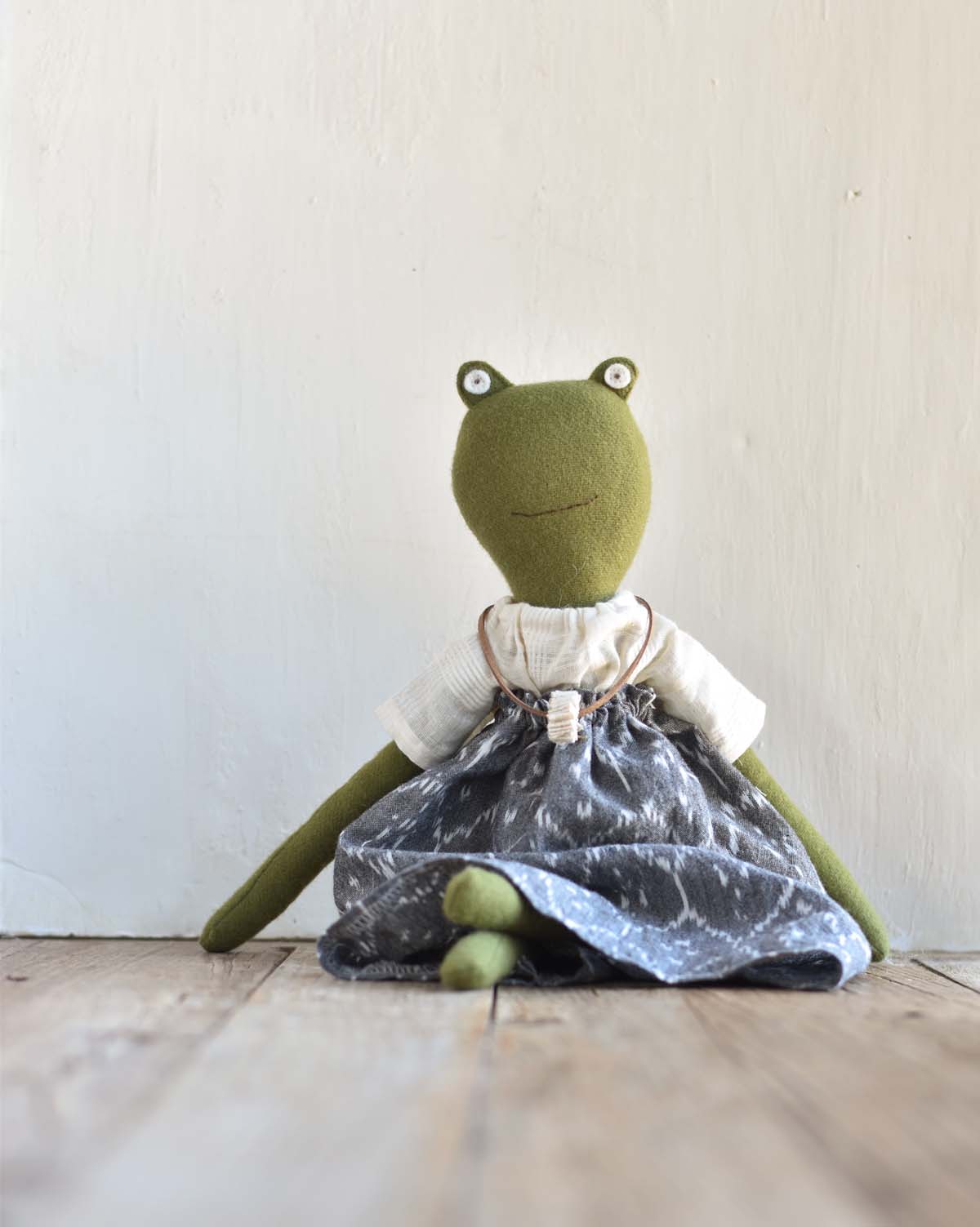 Fern the Frog, wearing an Ikat Field Skirt and white shirt, sits on a wooden floor against a white wall.