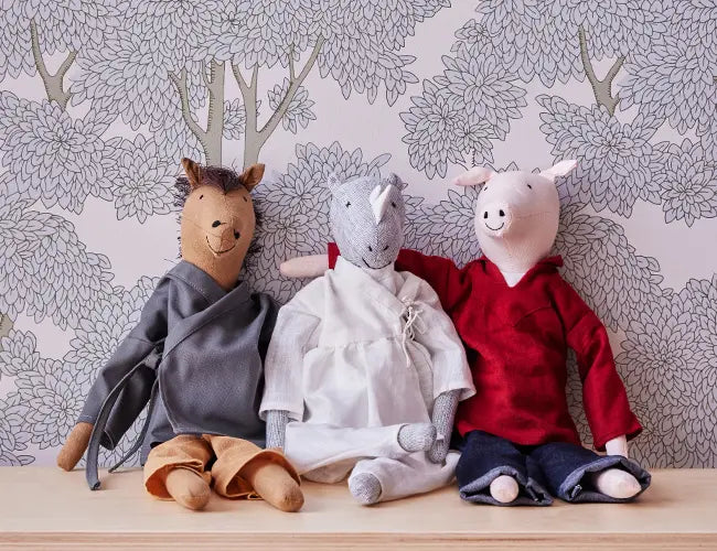 Three plush animals in robes: a deer with brown pants, a gray rhinoceros in white, and a pig in red, sit on a wooden surface against a floral wallpaper background.
