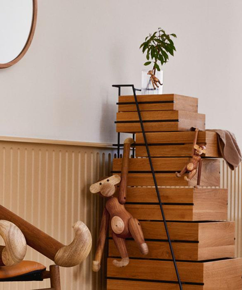 Stacked wooden drawers with a plant on top, Le Monkey by Kay Bojesen toys hanging, a chair, and a round mirror in a beige-paneled minimalist room.