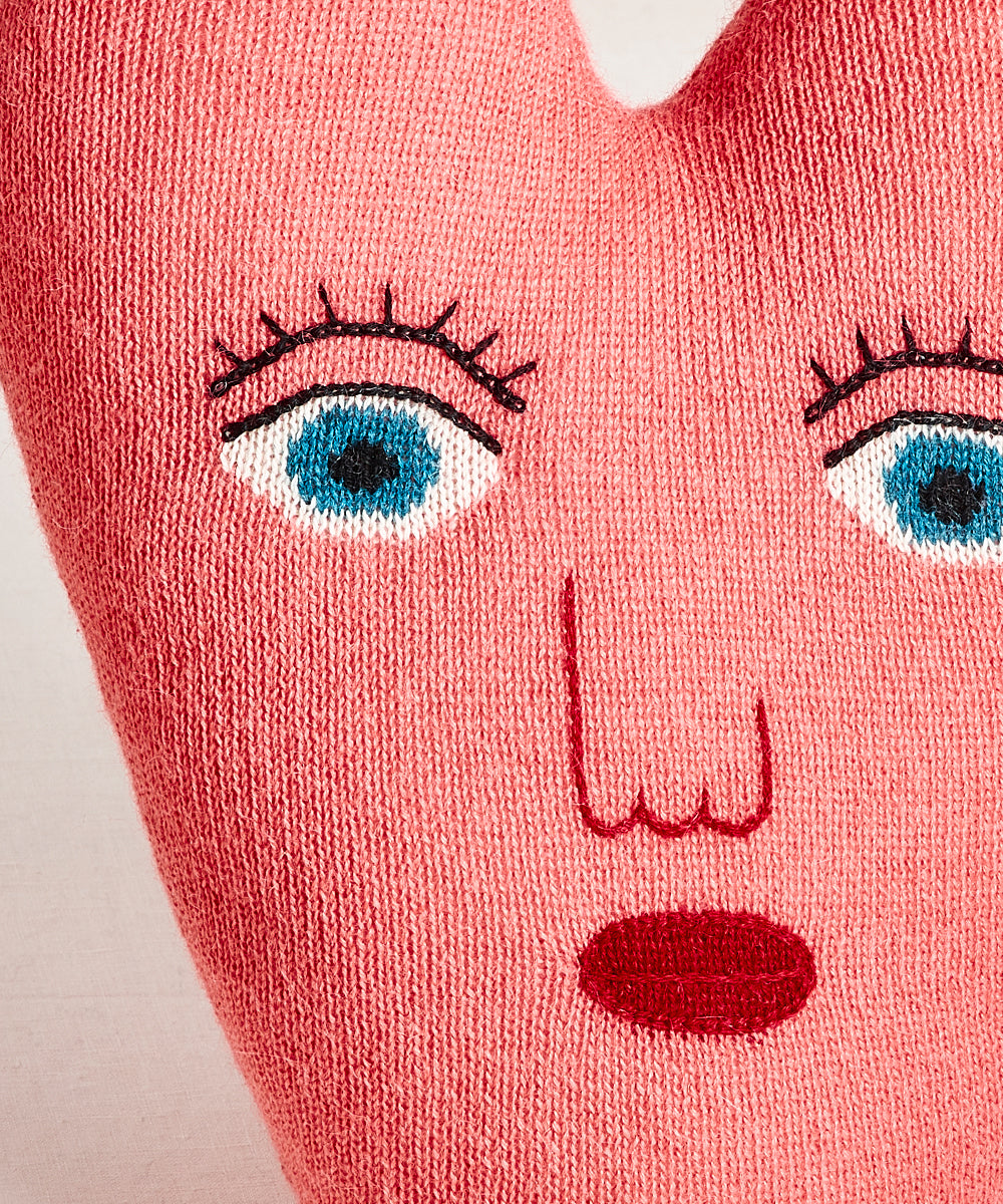The Heart Pillow is a pink knit cushion featuring embroidered blue eyes, eyelashes, nose, and red lips on a light background.