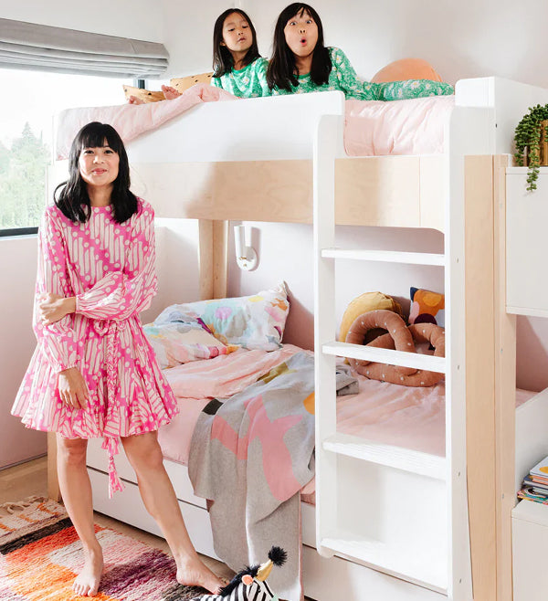 A woman in a pink dress stands by a white bunk bed with two children in green pajamas on the top bunk. The room has colorful decor and a window with a view of trees.