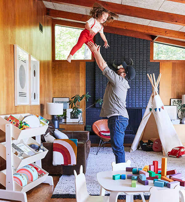 An adult playfully lifts a child in a mid-century modern living room with toys and a teepee.