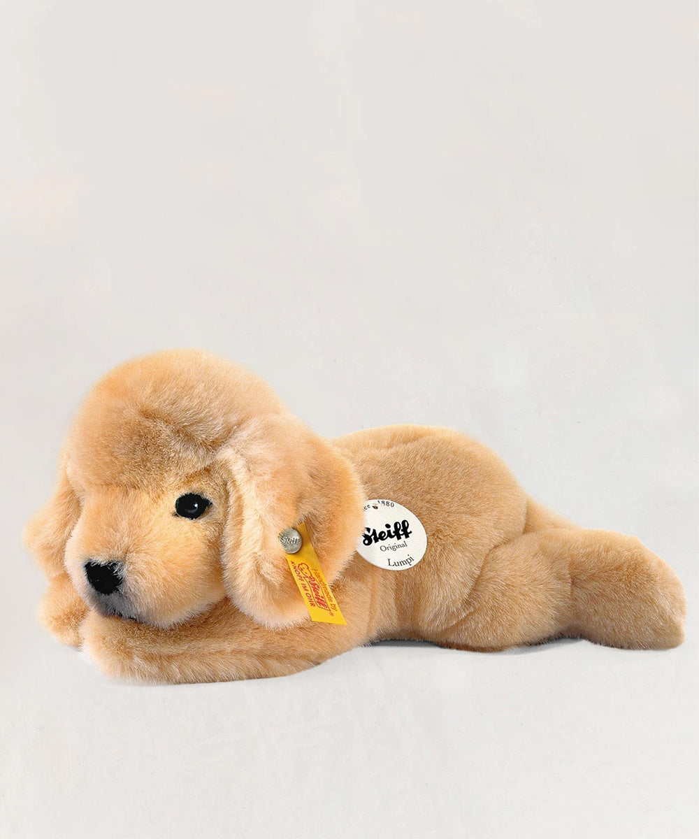 Le Puppy, a plush golden puppy toy from Steiff, lies down with brand tags on its ear and chest against a white background.