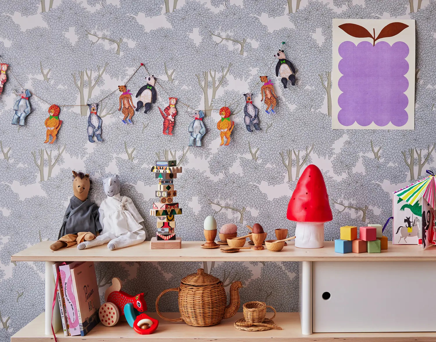 Childrens room with a shelf holding toys, books, and a red mushroom lamp. Animal garland hangs above, with tree-patterned wallpaper and a purple-framed artwork on the wall.