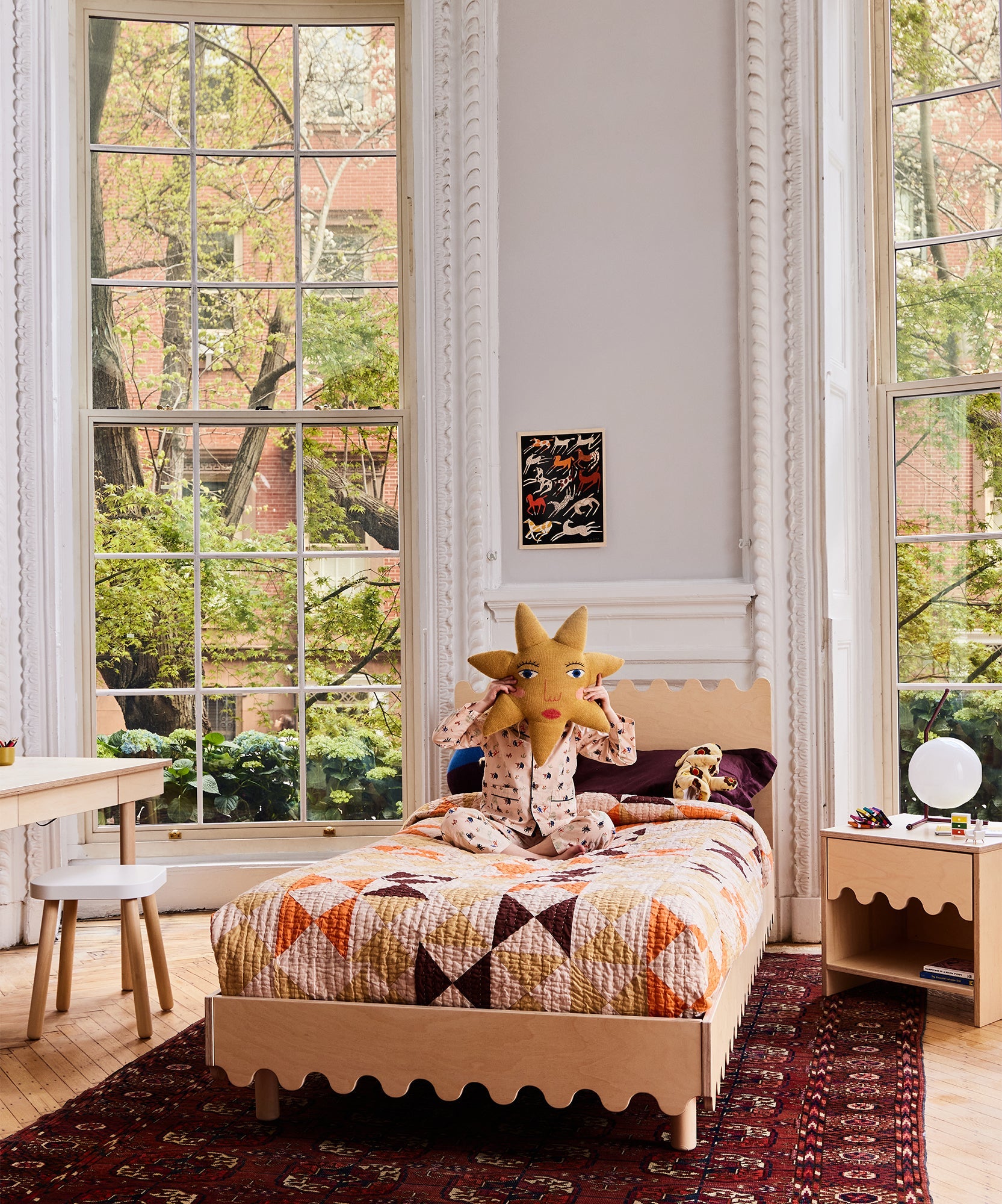 Child in patterned pajamas sits on a Moss Twin Bed, holding a star pillow in a bright bedroom with large windows.