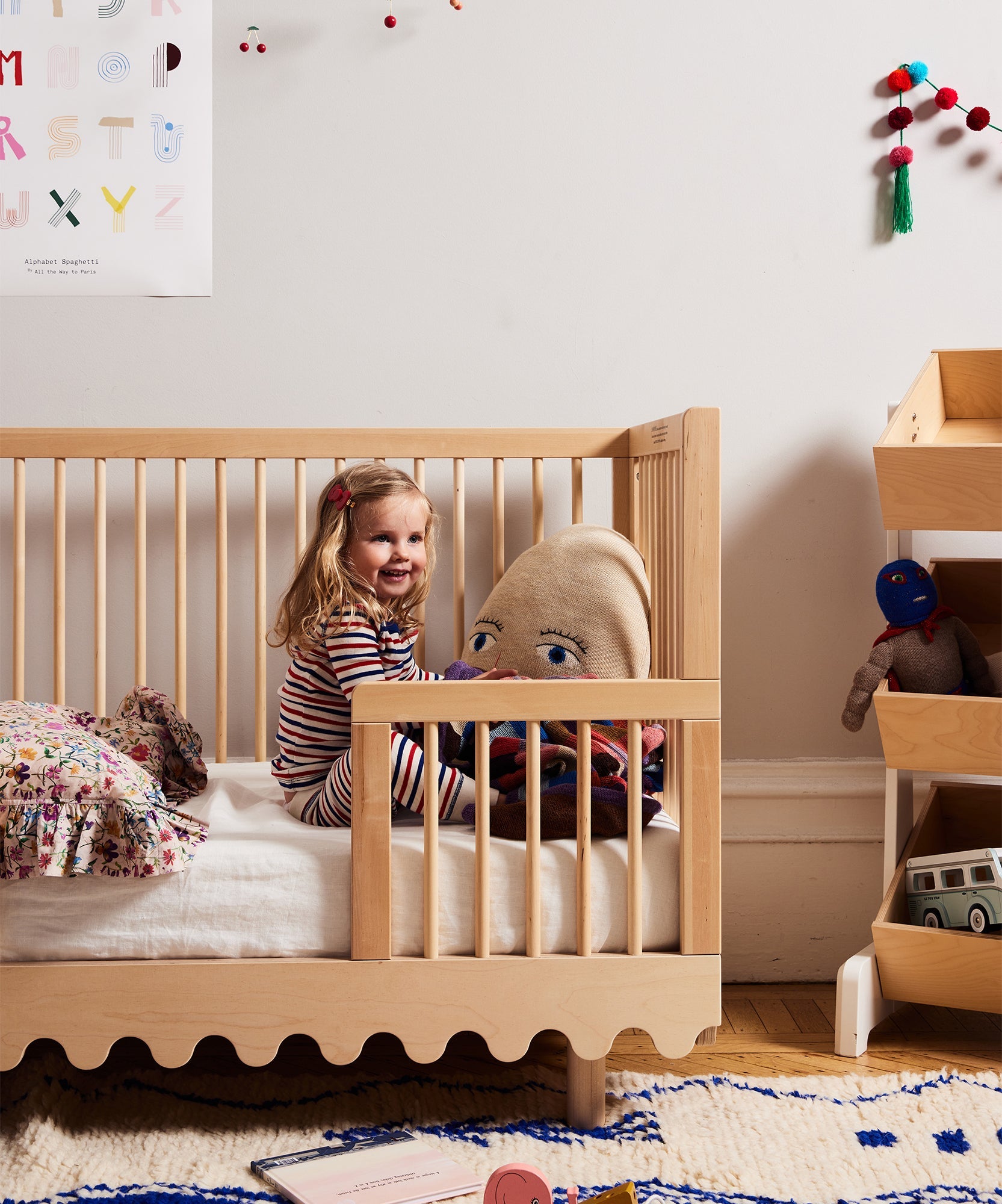 Smiling child in striped pajamas sits in a wooden crib featuring the Moss Crib Conversion Kit, cuddling a large plush toy in a cozy, decorated room.