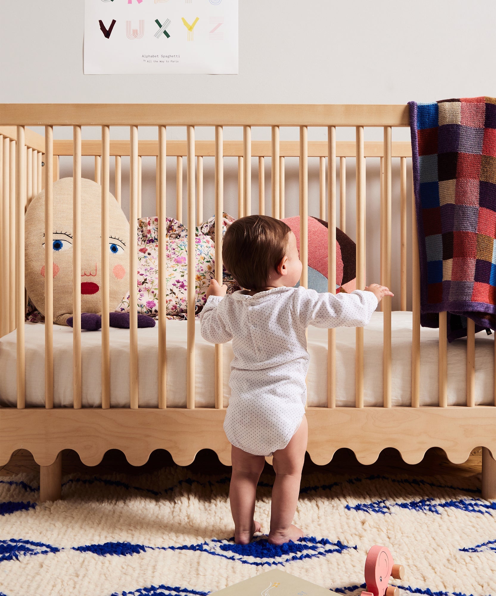A baby stands holding onto a Moss Crib, which contains colorful pillows and a plaid blanket.