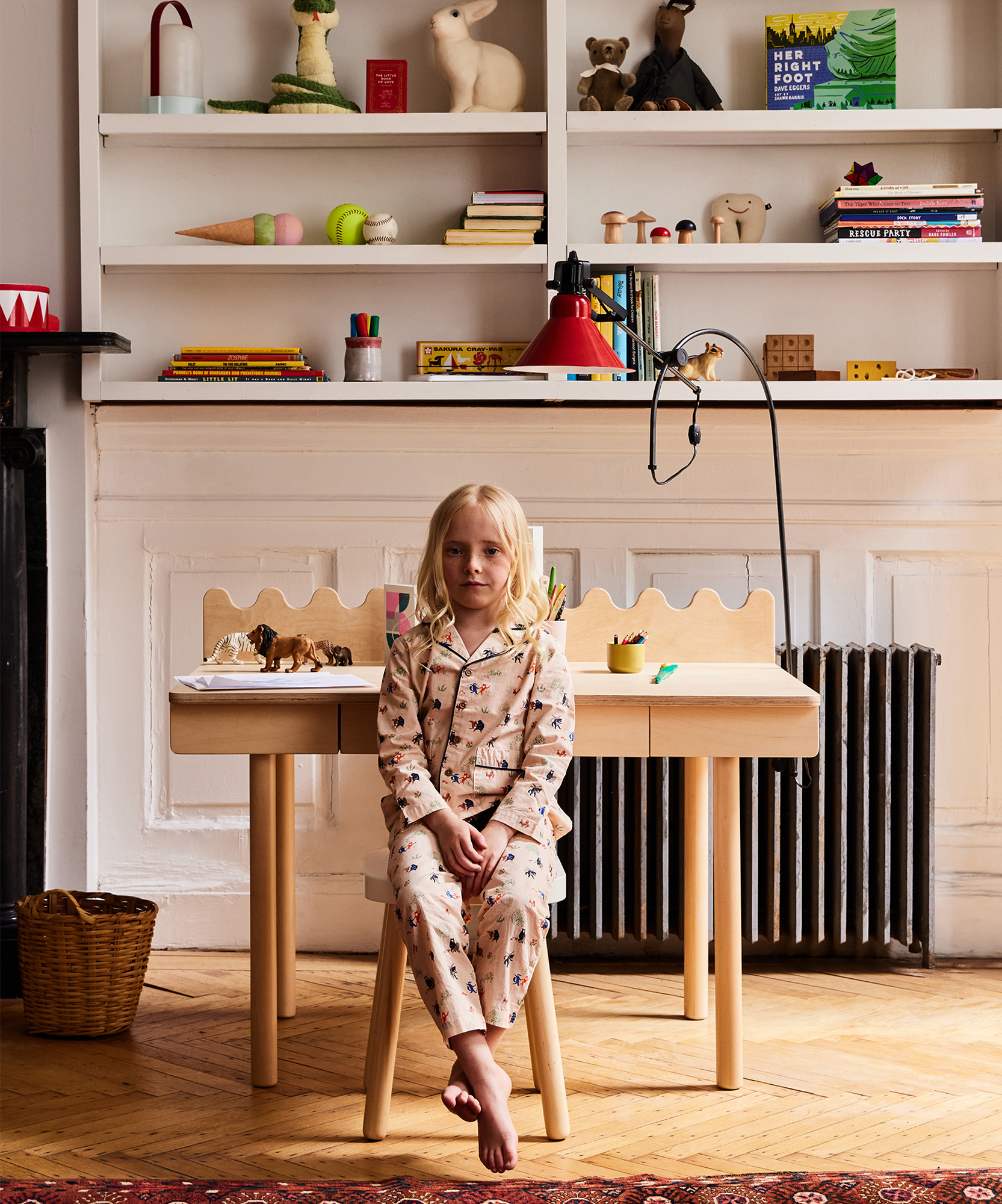 A child in pajamas sits at a Moss Desk in a cozy, colorful room filled with shelves and toys.