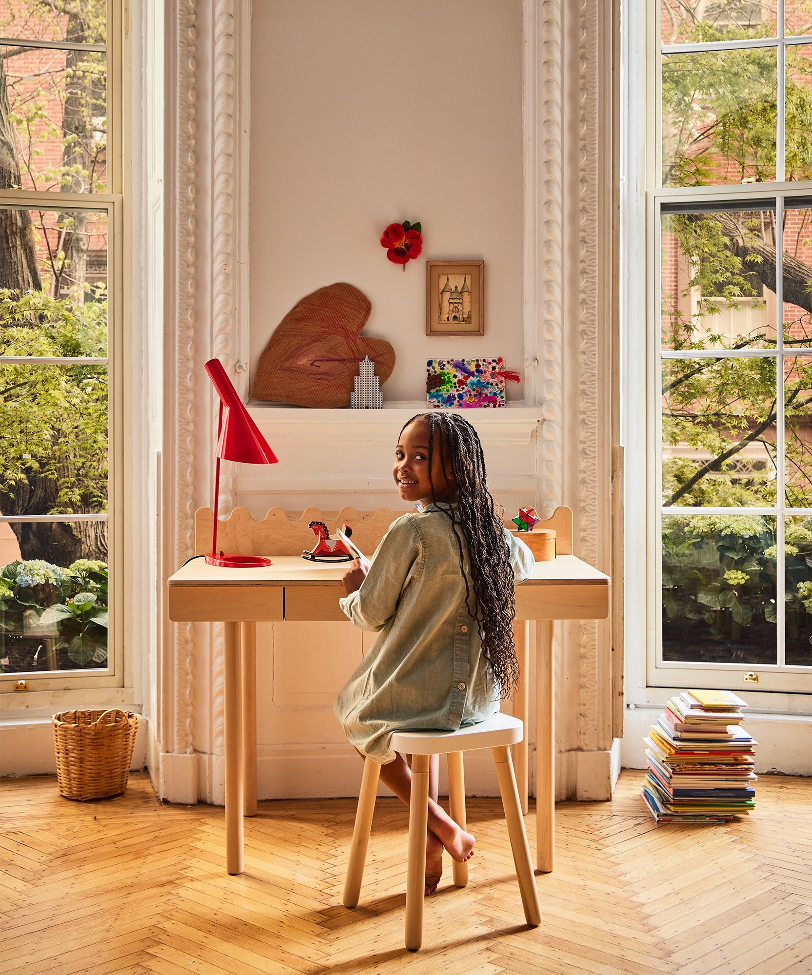 A girl smiles while sitting at a Moss Desk by tall windows, with books stacked on the floor nearby.