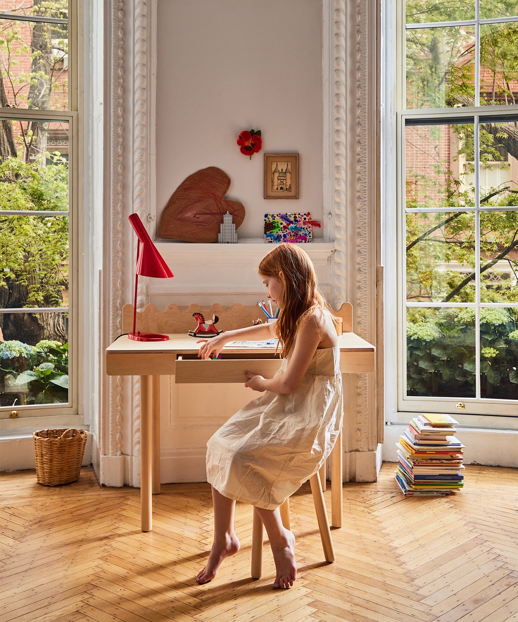 A young girl draws at a Moss Desk by large windows, with books stacked nearby and greenery visible outside.