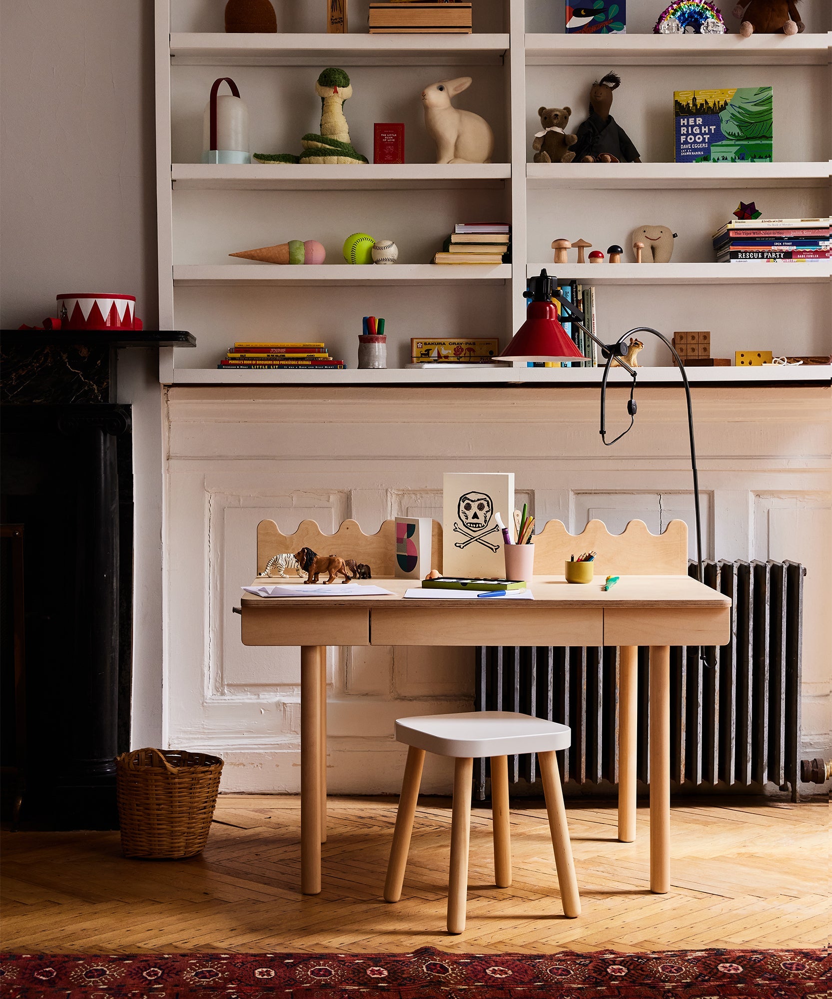 The Moss Desk for children shown with art supplies and toys, set before shelves filled with books and playful decor.