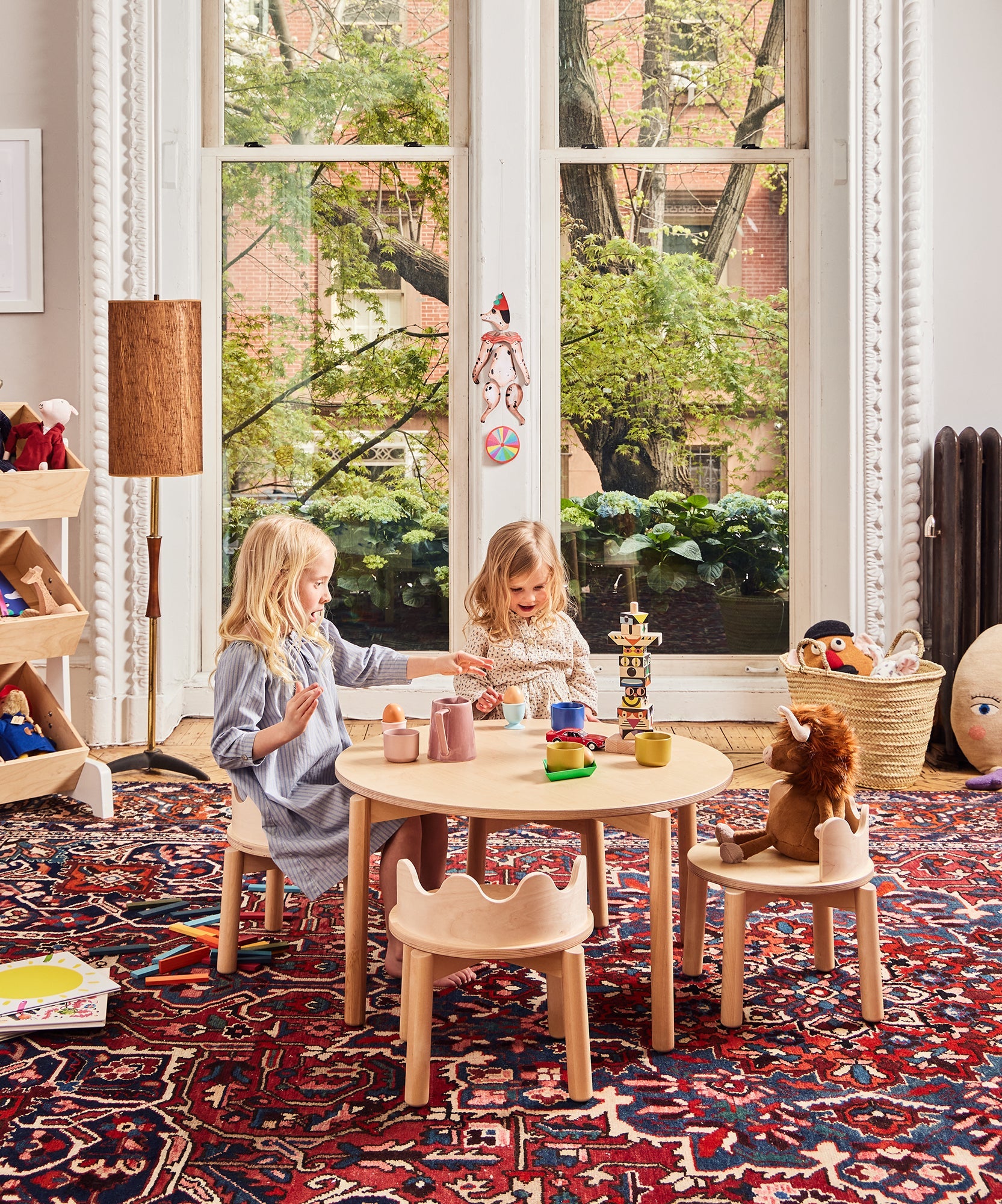 Two young girls sit on Moss Chairs (Set of 2) playing with toys at a small round table in a bright, cozy room with a large window and rug.