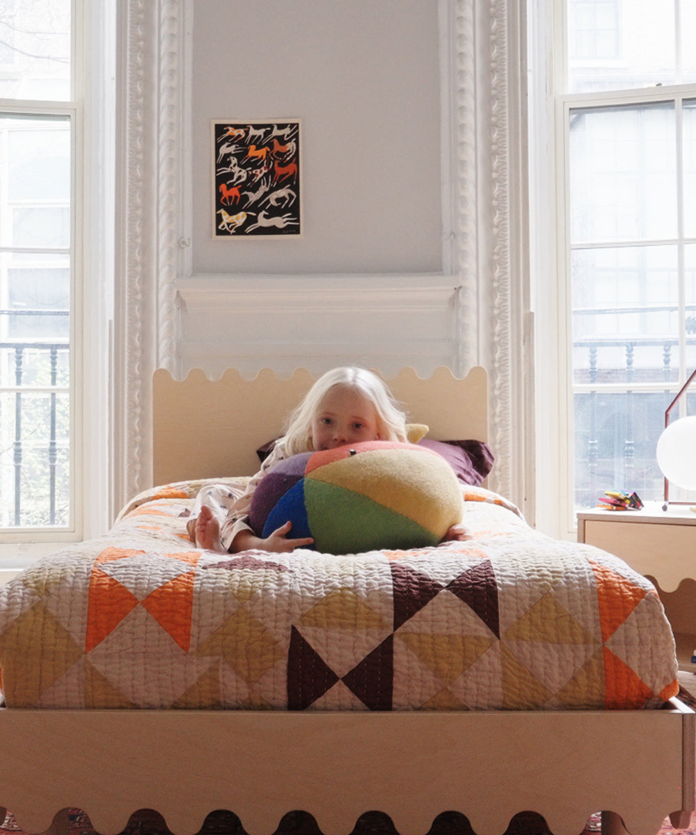 A child with light hair lies on a bed with a colorful quilt, holding the Circus Ball - Rainbow pillow.