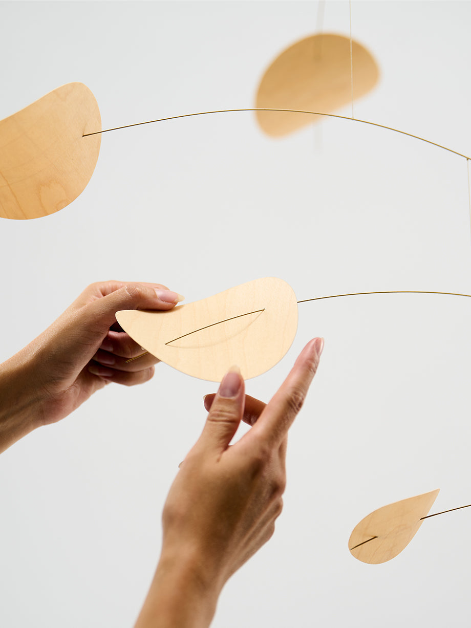 Two hands adjust the Drifting Clouds | Nature | Size M mobile, featuring leaf-shaped pieces hanging from thin wires against a white background.