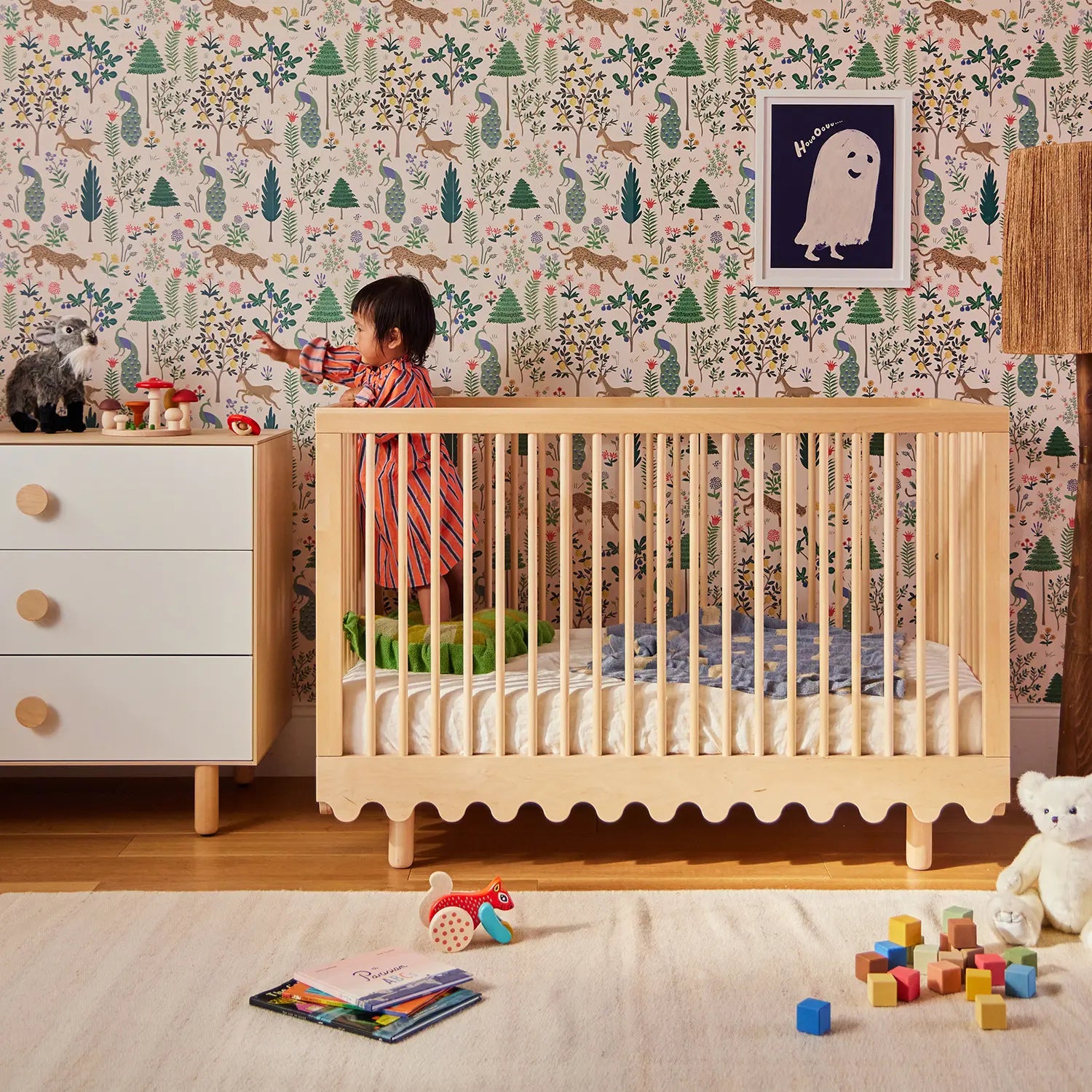 A toddler in a striped outfit stands in a crib, reaching for stuffed animals on a dresser. The room has patterned wallpaper, a rug, colorful blocks, and a toy train on the floor.