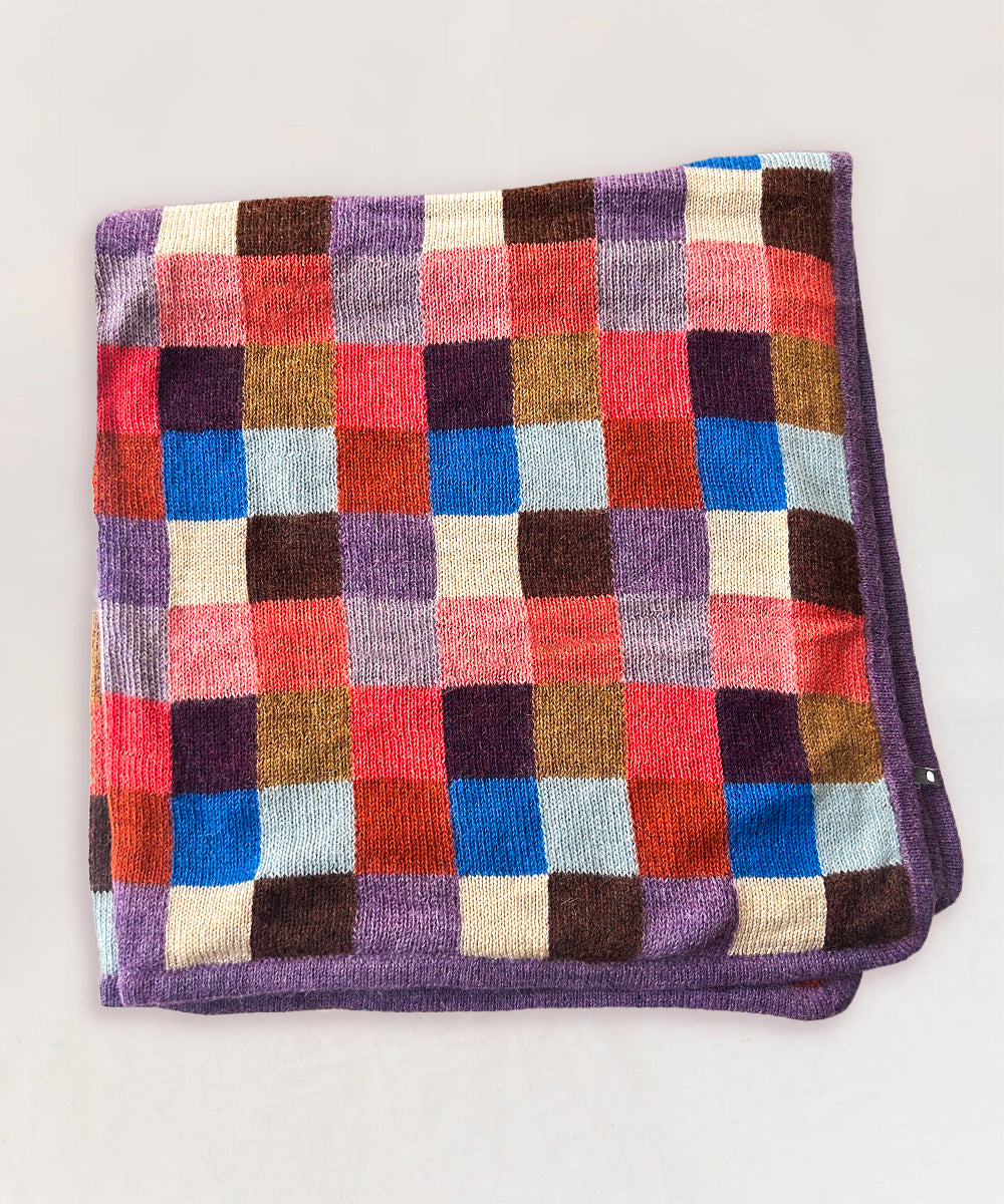 The Patchwork Blanket is a multicolored checkered knit throw with purple edges, shown folded on a light background.
