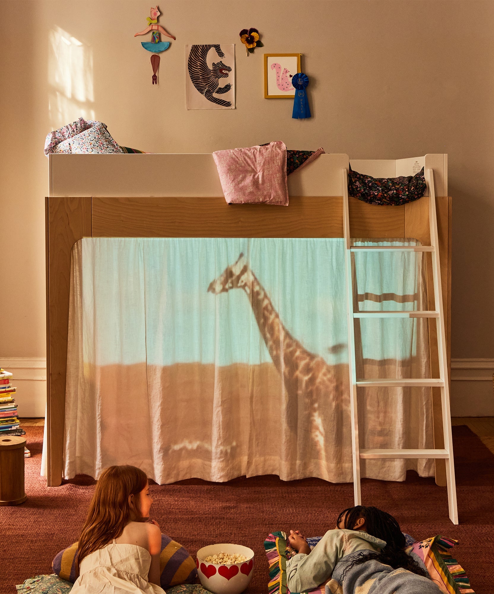 Two children watch a giraffe projected onto Perch Bunk Bed Curtains beneath a loft bed decorated with art and pillows.