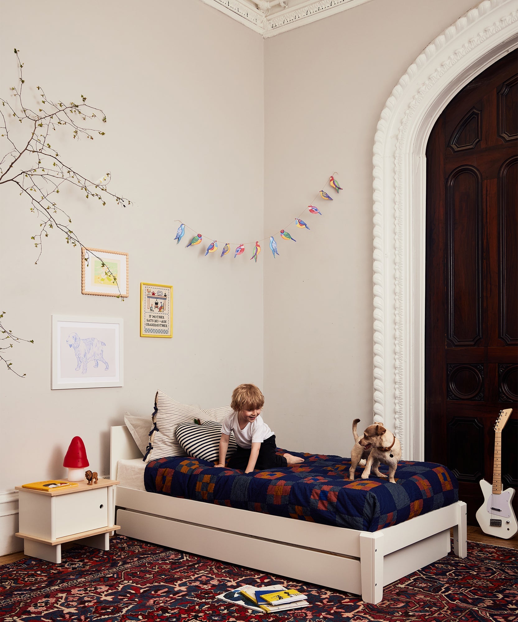 A child sits on a Perch Twin Bed with a toy dog in a cozy, decorated bedroom featuring art, books, and a guitar.