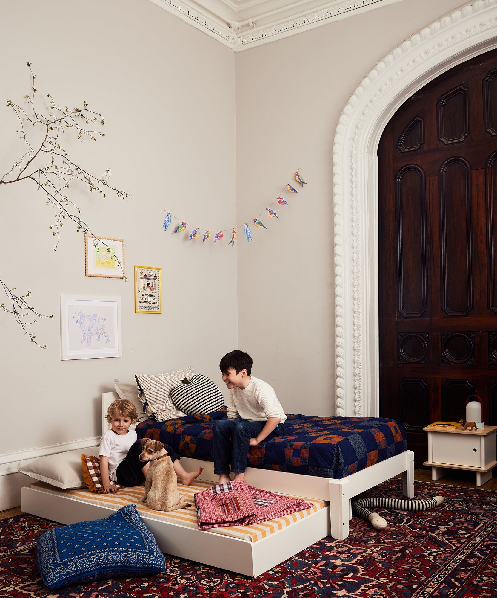 Two children and a dog relax on Perch Twin Beds in a cozy, decorated bedroom with patterned rugs and wall art.