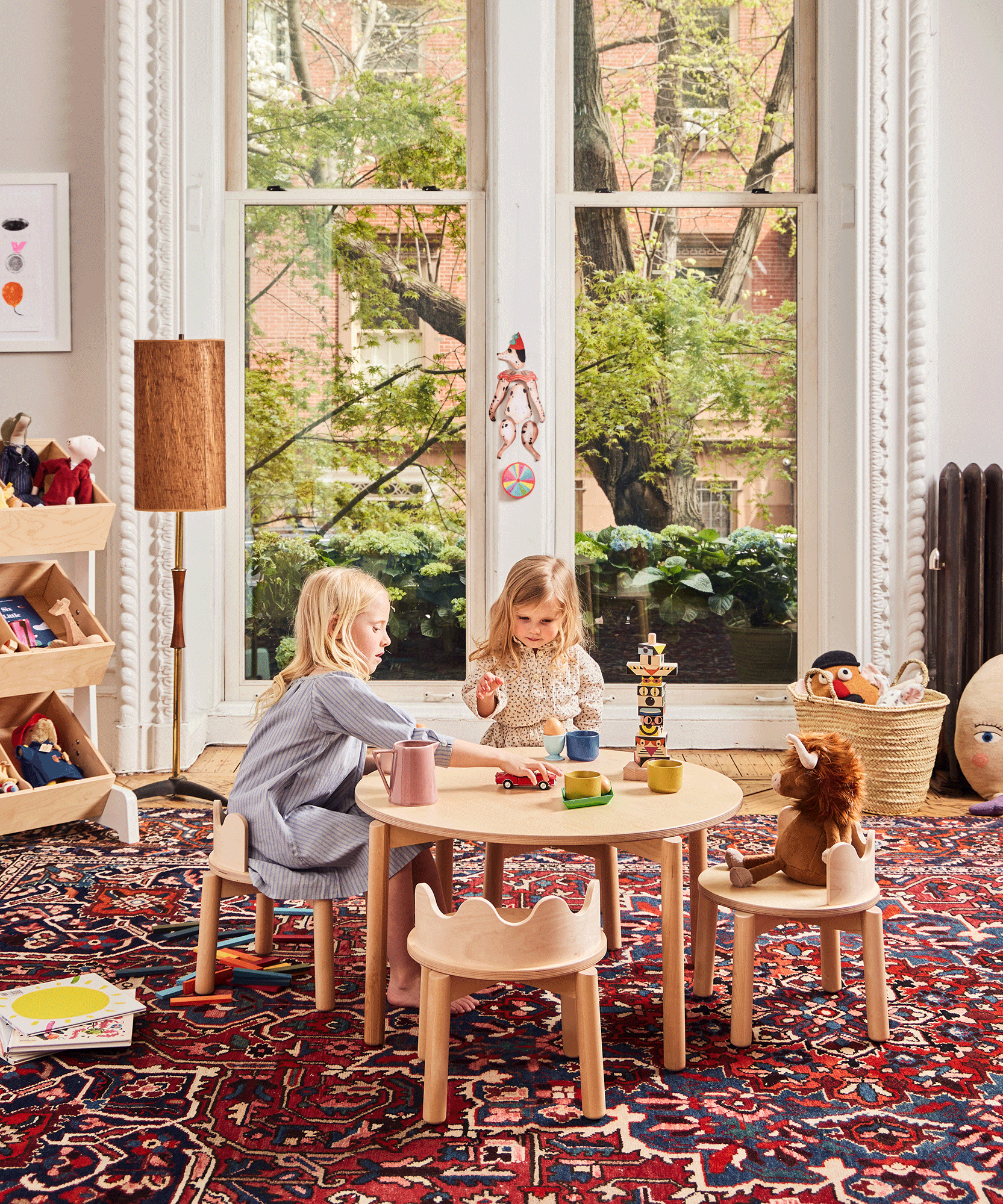 Two young children play at a Moss Table in a cozy, sunlit room with large windows and a patterned rug.