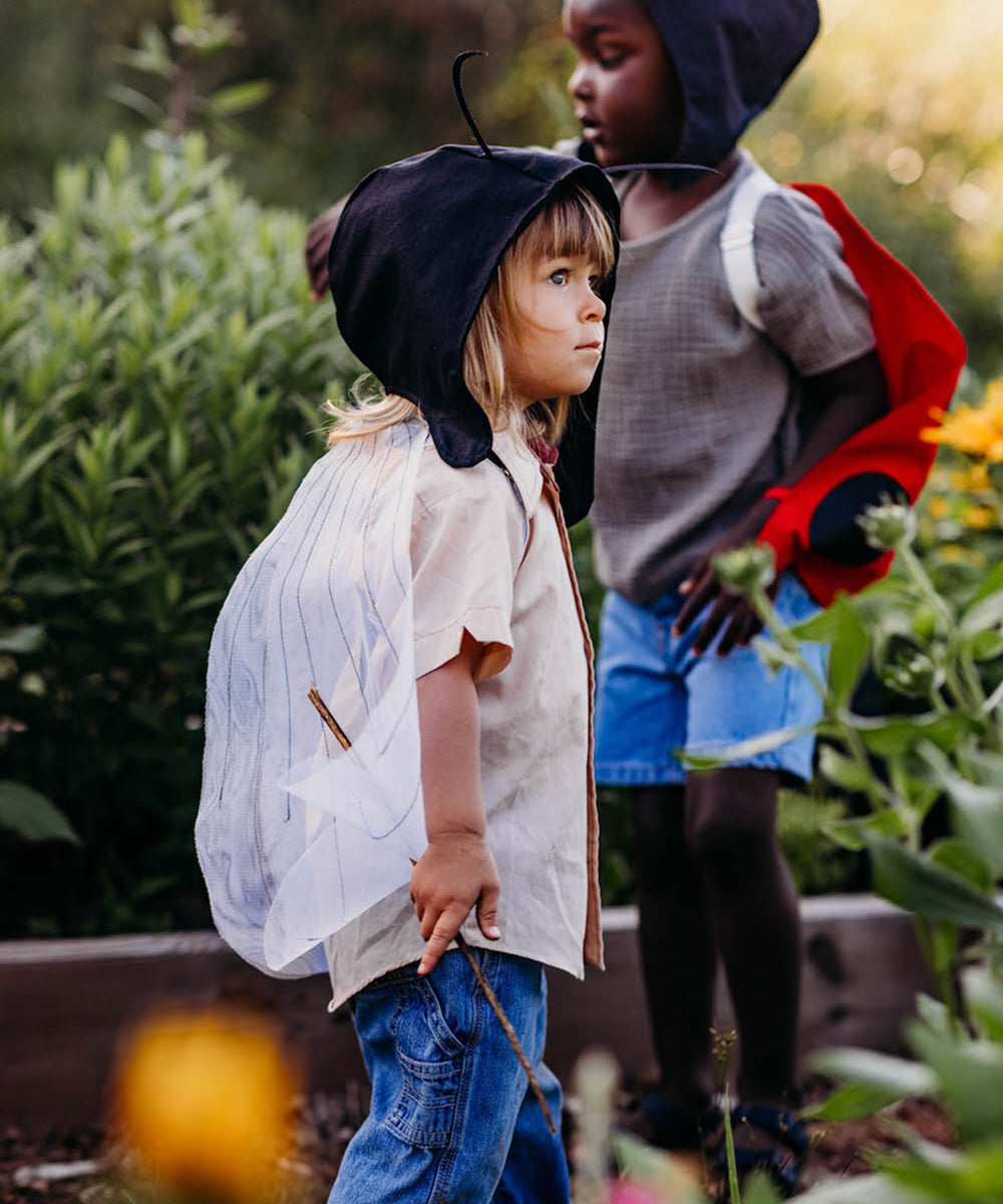Two children play outdoors; one wears Bumble Bee Costume Wings with antennae, the other a red cape and hood. They’re surrounded by green plants.