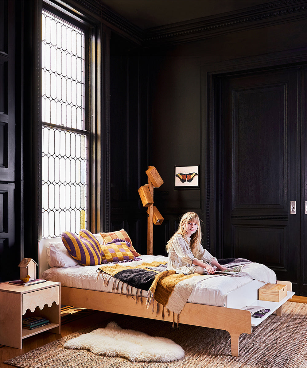 A woman sits on a stylish bed featuring the Naturepedic 2-in-1 Organic Waterproof Mattress in a chic, dark-walled room with large windows and modern decor.