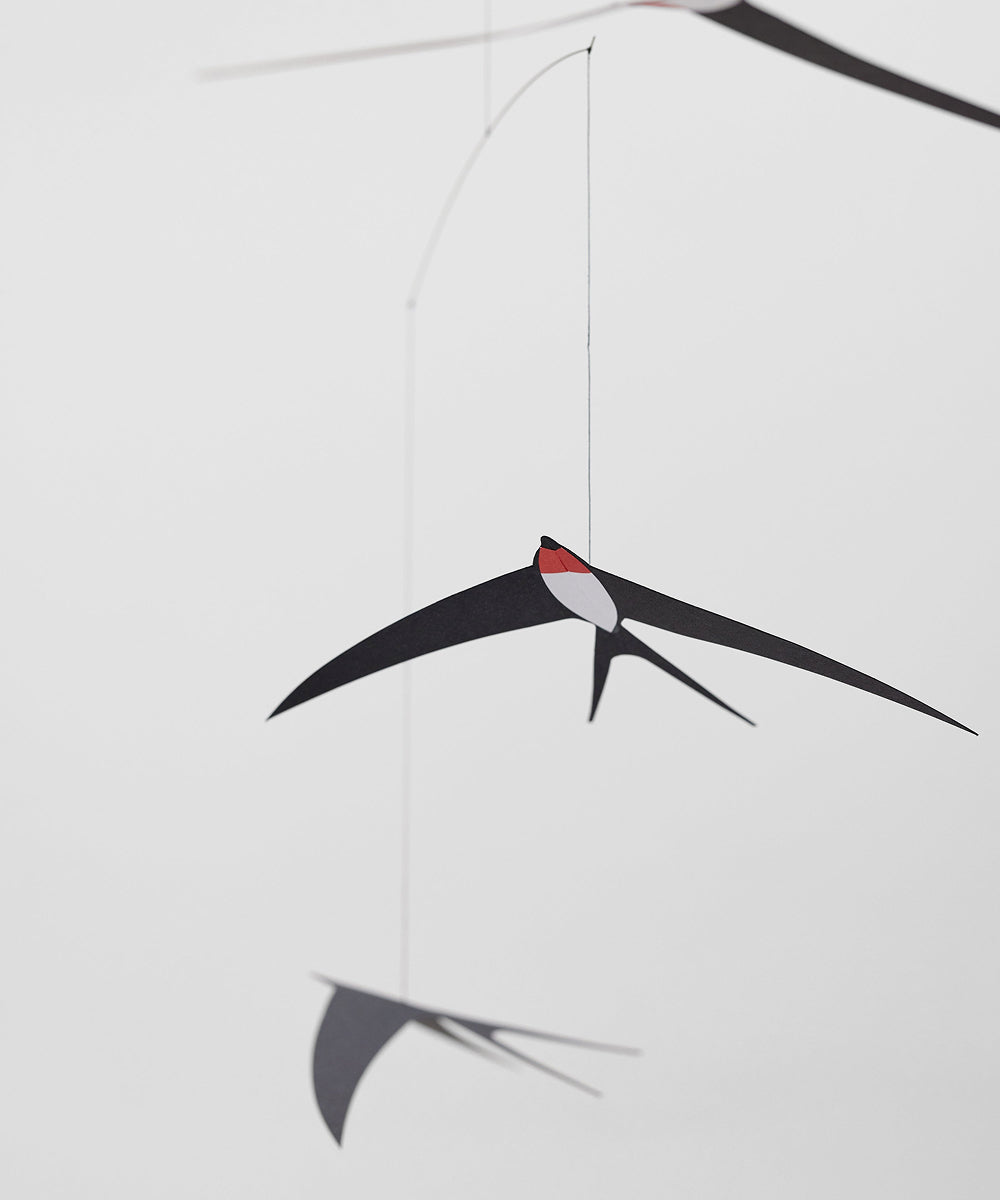 Le Mobile - Flying Swallows: Minimalist mobile with paper swallows, one with a red head spot, suspended by thin strings on a plain white background.