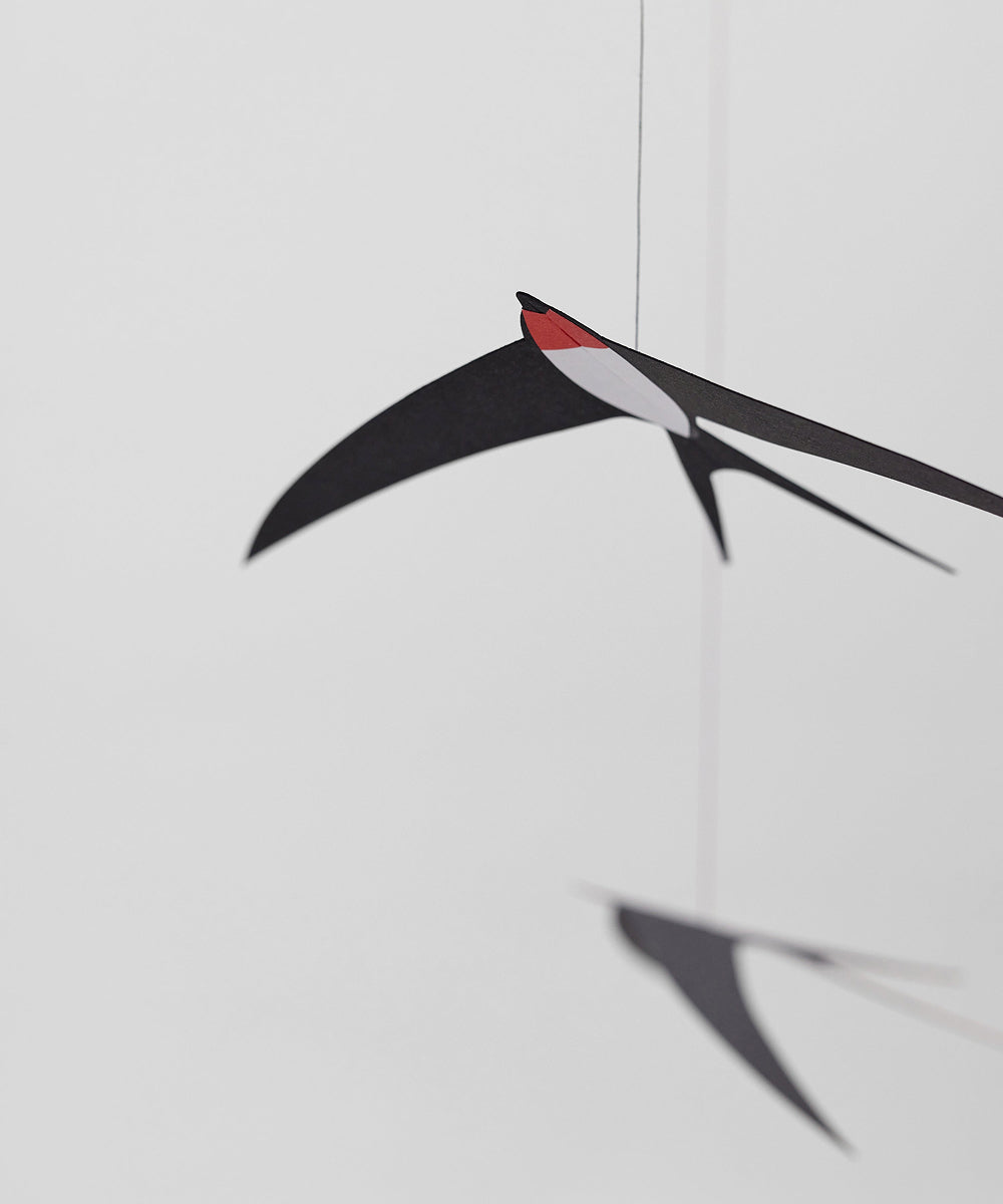 Le Mobile - Flying Swallows: Minimalist mobile of black paper swallows with red faces, suspended by thin strings against a plain white background.