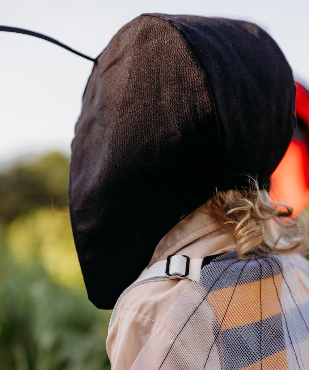 Person in a Le Antennae Hat and ant costume with mesh wings, seen from the side outdoors, greenery in background.