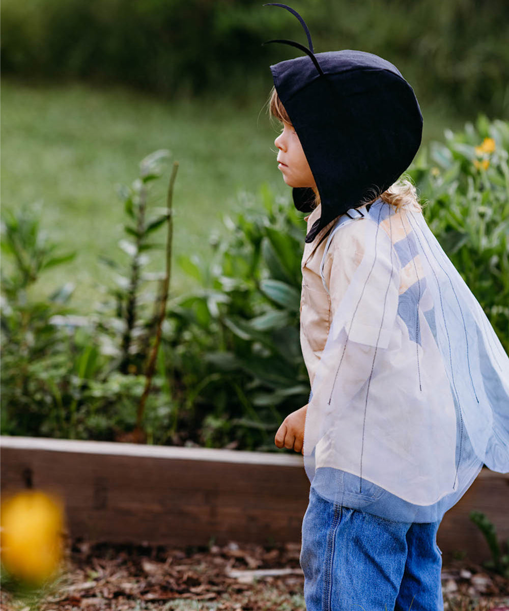 A child in a Le Antennae Hat stands outdoors near plants, wearing wings and looking to the right.