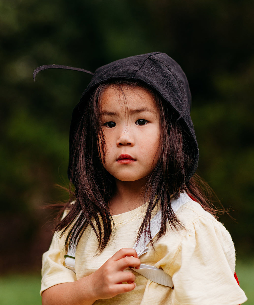 A young girl with long dark hair wears the Le Antennae Hat and a yellow shirt, looking seriously at the camera outdoors.