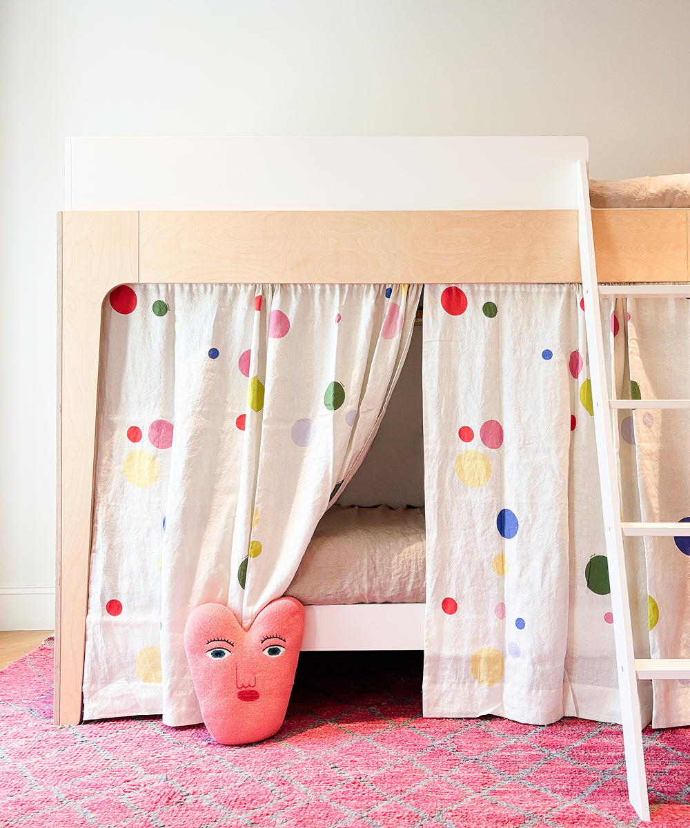 Printed Perch Bunk/Loft Linen Curtains with a colorful polka dot print hanging from the bottom bunk of a wooden bed frame, with a heart-shaped face pillow on the floor.
