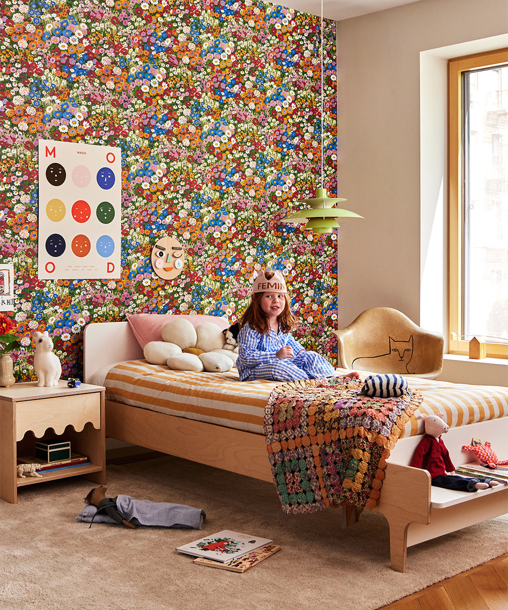 Child with a Daisy Pillow in striped bed, surrounded by colorful floral wallpaper and eclectic decor. Posters on the wall, window on right letting light in.