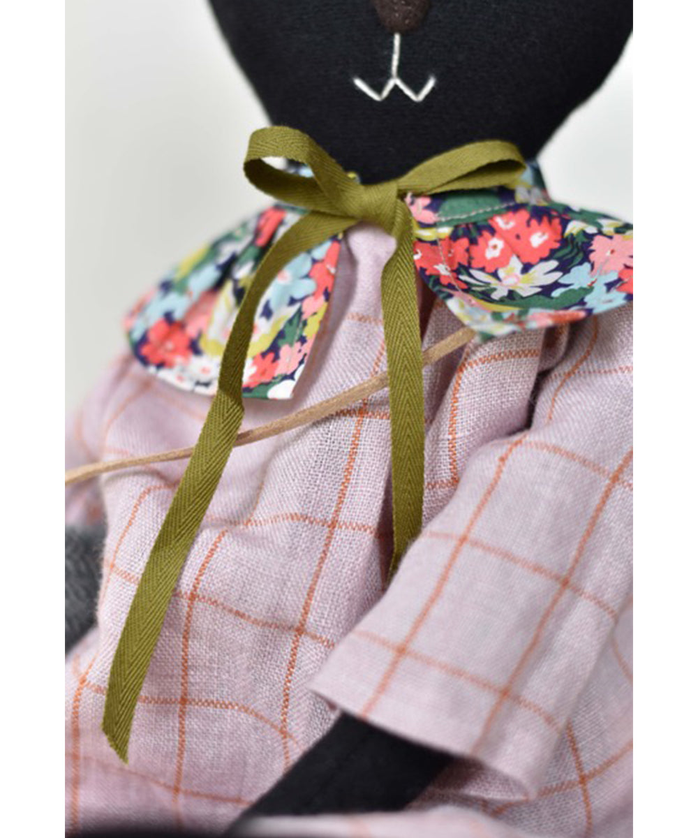 Scout le Skunk wears a plaid pink dress with a floral collar and a green ribbon bow.