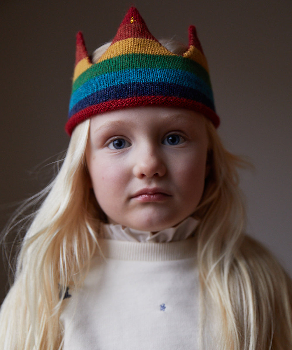 A child with long blonde hair wears a cream sweater and the Rainbow Crown, looking at the camera.