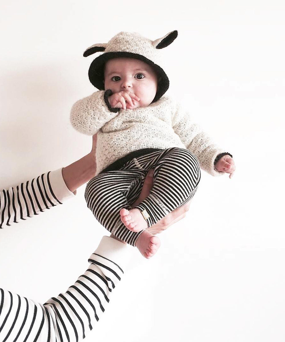 A baby wearing the Sheep Hoodie with animal ears and striped pants is held up by an adult in a striped shirt.