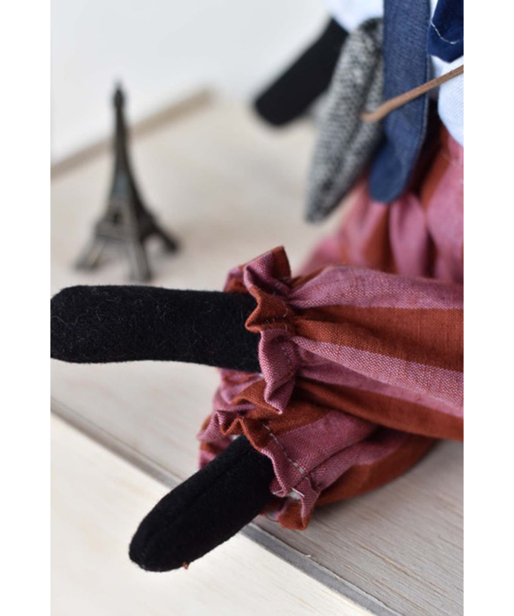 Theo le Skunk, a fabric doll with black legs and ruffled red pants, sits near a small Eiffel Tower figurine.