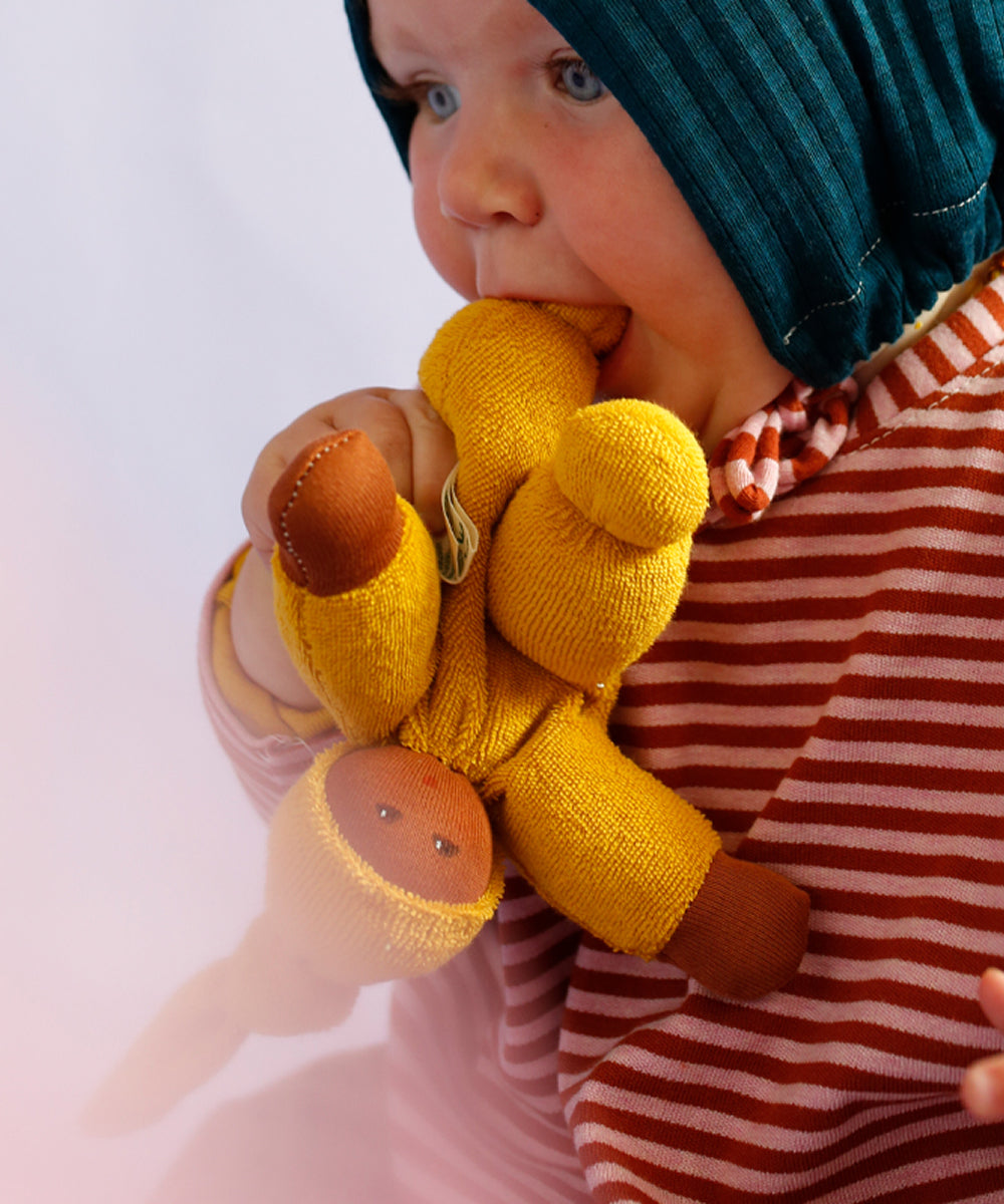 Baby in striped shirt and blue bonnet biting a Waldorf Doll - Blue with yellow body and brown hands and feet.