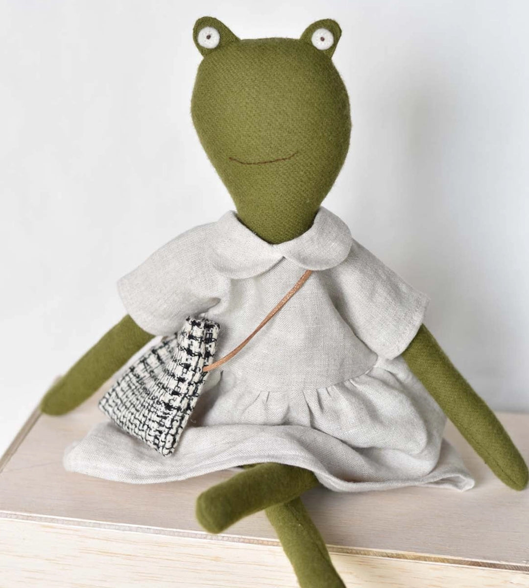 Fern le Frog, dressed in a light outfit, sits with crossed legs and holds a small bag.