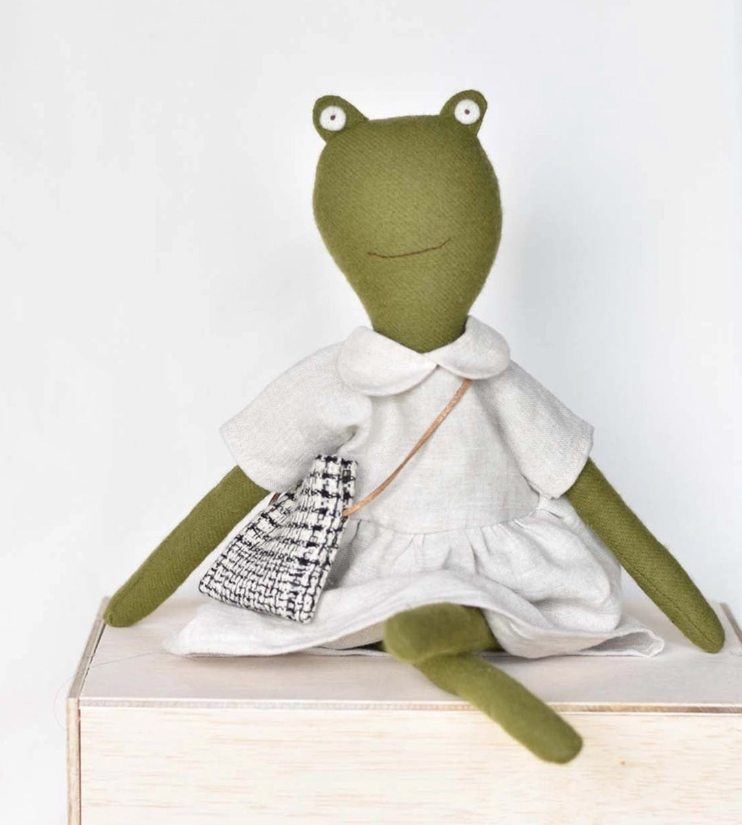 Fern le Frog is a green plush toy in a white dress with a patterned bag, sitting on a wooden box.